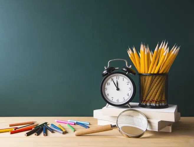 Na biurku po lewej stronie leżą kredki, po prawej stronie trzy książki, na książkach zegar oraz pojemnik, w pojemniku ołówki