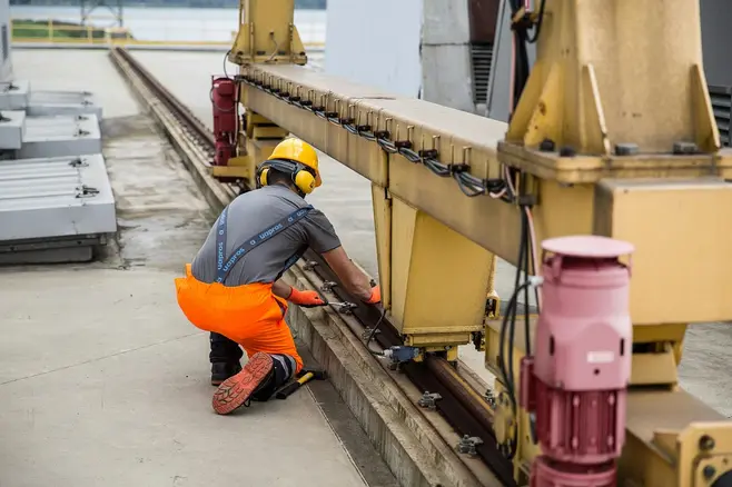 Man in work clothes, wearing a helmet and ear protection, tightening bolts on a machine with a wrench